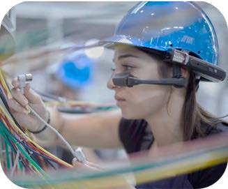 A female frontline worker wearing a blue hard hat and a wearable AR glasses headset while inspecting complex electrical wiring, demonstrating augmented intelligence and human-AI collaboration in an industrial environment.